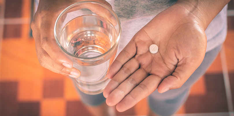 Round pill held in one hand and glass of water held in the other